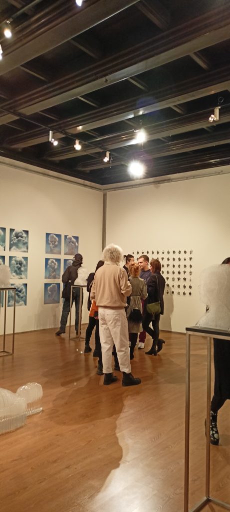 ludzie w galerii, w tle obrazy niebiesko-białe, chmury, abstrakcyjne twarze